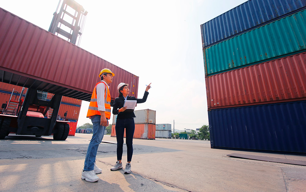Agenciamento de cargas de importação e/ou exportação em porto, com homem em mulher com roupas de trabalho apontando para cargas