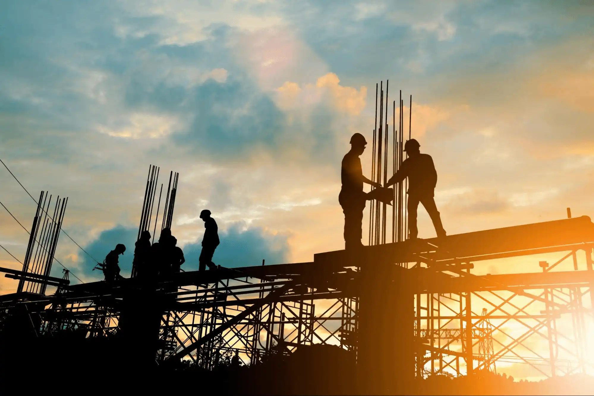 Trabalhadores da construção civil em uma obra ao pôr do sol, montando estrutura metálica.